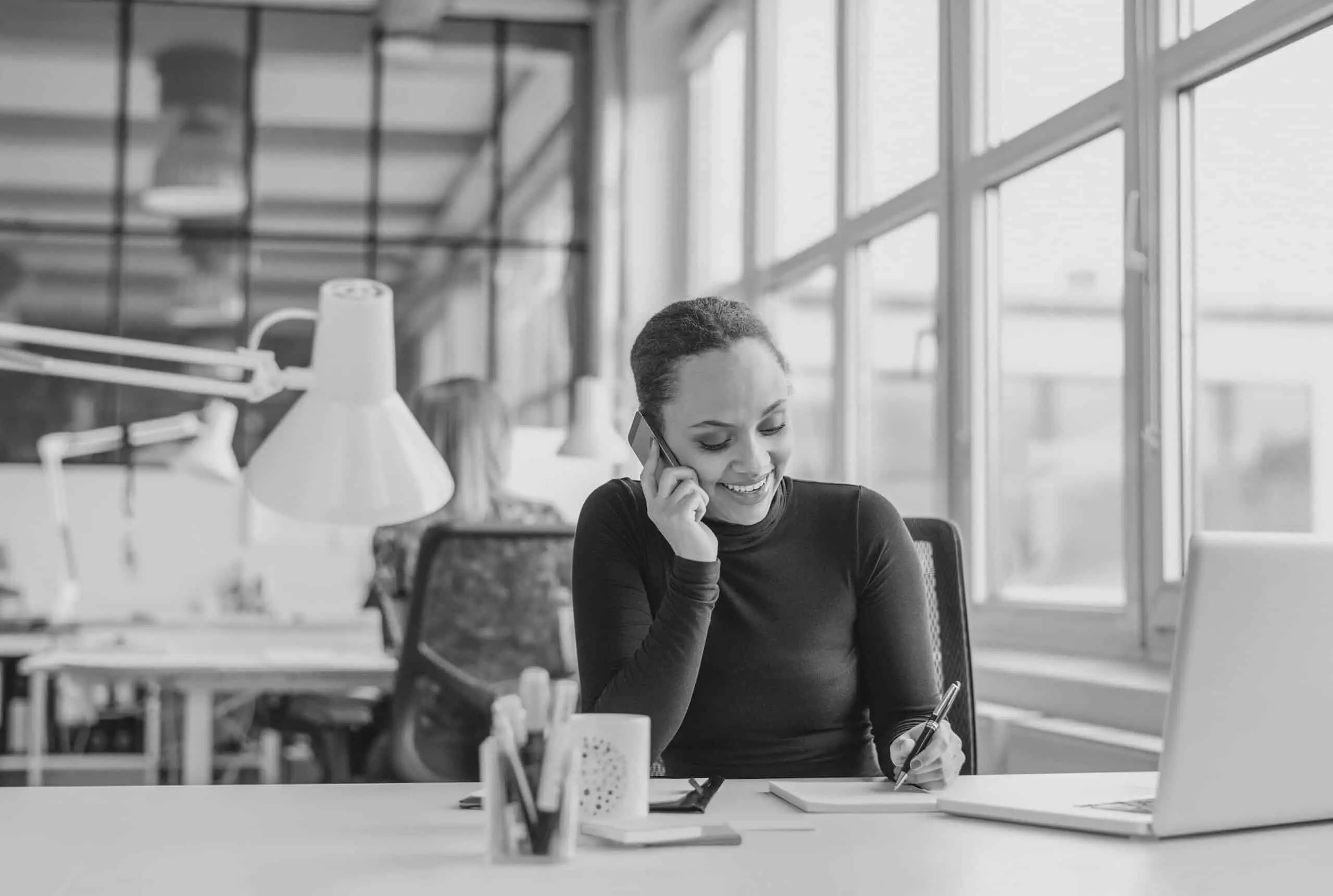 Happy young woman taking notes while talking on mobile phone. African woman working at her desk answering a phone call.