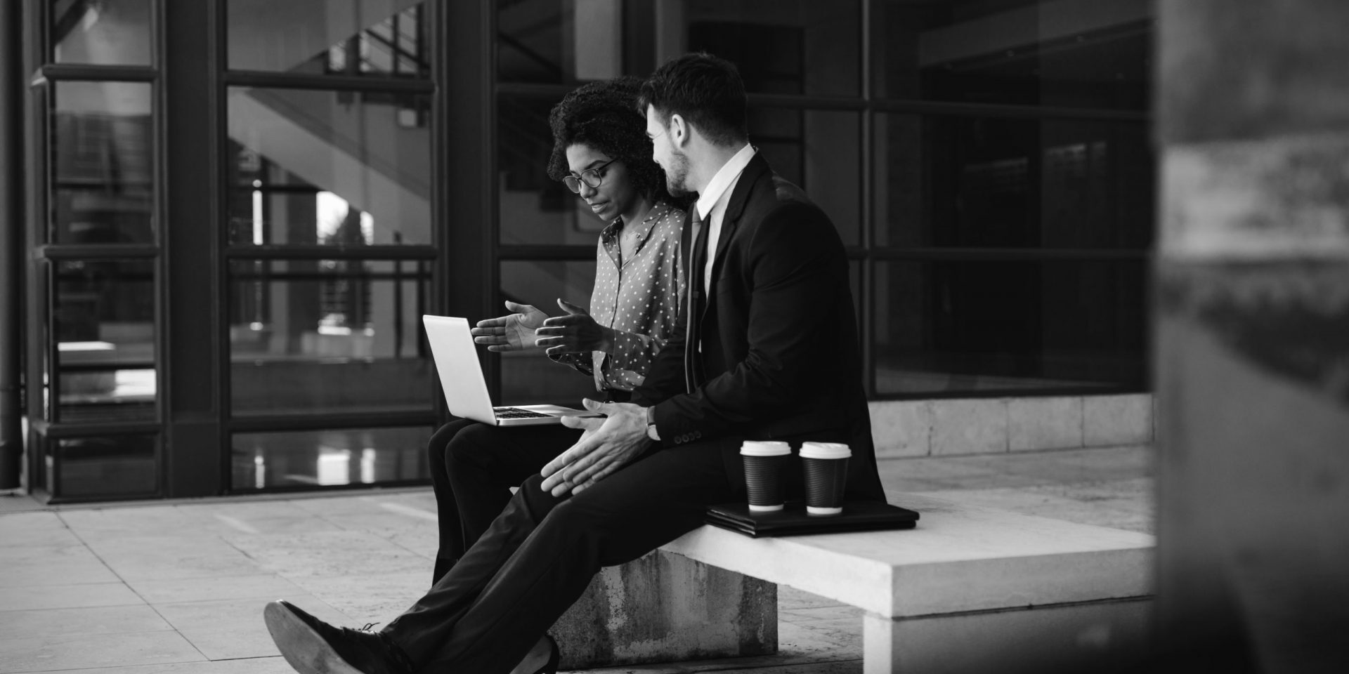 Two business people sitting on a bench in modern office building with a laptop. Woman with laptop discussing business with male colleague.