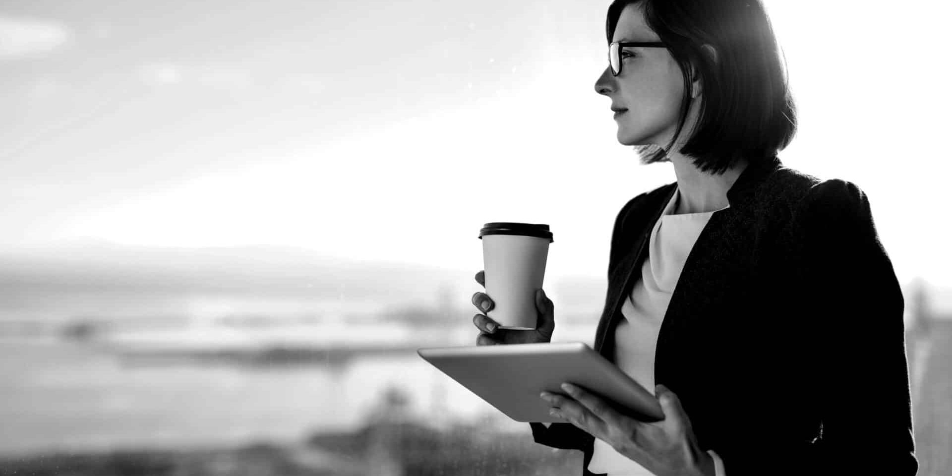 Attractive businesswoman drinking a coffee and using a digital tablet while standing at a window in an office overlooking the city